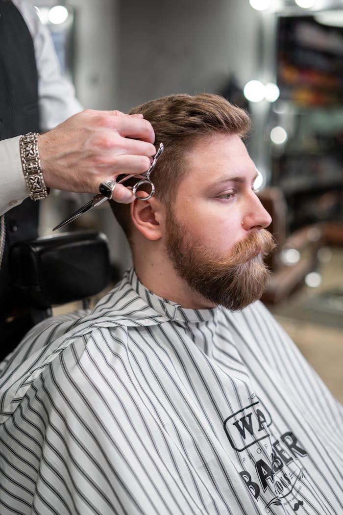 A barber trims a bearded man's hair in a classic barbershop setting.