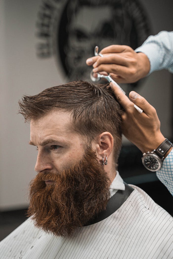 Close-up of a barber styling a bearded man's hair indoors. Side view capturing the precise haircut process.